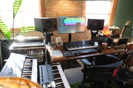 A home music studio featuring multiple electronic keyboards, a large monitor displaying audio editing software, and studio speakers. The room has a cozy vibe with a brick wall, wooden desk, potted plant, and a salt lamp. Various musical and audio equipment are neatly arranged, including synthesizers and mixers, alongside a comfortable office chair.