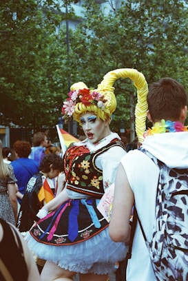 A person dressed in elaborate, colorful clothing and makeup is captured in a vibrant outdoor setting. They wear a floral headdress and an intricately designed outfit with a tutu, surrounded by a crowd of people, some adorned with rainbow accessories. Lush green trees form the background, and a rainbow flag is partially visible.