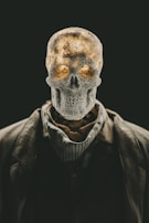 A human skeleton head with textured surface placed atop a body dressed in a suit and scarf. The image has a dramatic contrast with a dark background, highlighting the skeletal structure. The lighting gives a warm glow to parts of the skull, enhancing its eerie and mysterious appearance.