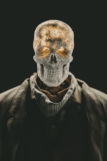 A human skeleton head with textured surface placed atop a body dressed in a suit and scarf. The image has a dramatic contrast with a dark background, highlighting the skeletal structure. The lighting gives a warm glow to parts of the skull, enhancing its eerie and mysterious appearance.