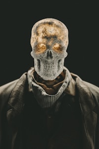 A human skeleton head with textured surface placed atop a body dressed in a suit and scarf. The image has a dramatic contrast with a dark background, highlighting the skeletal structure. The lighting gives a warm glow to parts of the skull, enhancing its eerie and mysterious appearance.
