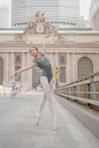 A ballet dancer in a gray leotard and white tights performs an elegant pose on a city street. The background features a historic building with a clock and intricate architectural details. A child on a bicycle is visible in the distance, along with several pedestrians.