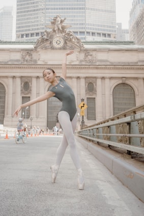 A ballet dancer in a gray leotard and white tights performs an elegant pose on a city street. The background features a historic building with a clock and intricate architectural details. A child on a bicycle is visible in the distance, along with several pedestrians.