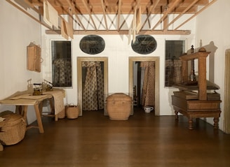A small room with wooden flooring features a collection of woven baskets, a wooden table, and a wooden drying rack on the ceiling. The room is decorated with patterned curtains and textiles. There are two black irons on the table, and a wooden press is located on the right side of the room. Light-colored textiles hang from the drying rack.