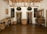 A small room with wooden flooring features a collection of woven baskets, a wooden table, and a wooden drying rack on the ceiling. The room is decorated with patterned curtains and textiles. There are two black irons on the table, and a wooden press is located on the right side of the room. Light-colored textiles hang from the drying rack.
