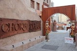 A rustic entrance marked by a large wooden sign displaying the name 'CHOKHI DHANI.' The entrance is flanked by two colorful, traditional statues dressed in ornate outfits. Beyond the archway, there's a statue of a woman and a sign indicating a restricted area for authorized personnel only. The surrounding architecture features earthy tones and intricate designs, typical of traditional Indian styles.