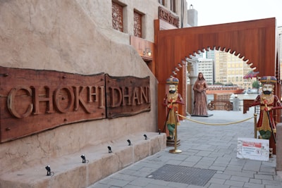 A rustic entrance marked by a large wooden sign displaying the name 'CHOKHI DHANI.' The entrance is flanked by two colorful, traditional statues dressed in ornate outfits. Beyond the archway, there's a statue of a woman and a sign indicating a restricted area for authorized personnel only. The surrounding architecture features earthy tones and intricate designs, typical of traditional Indian styles.