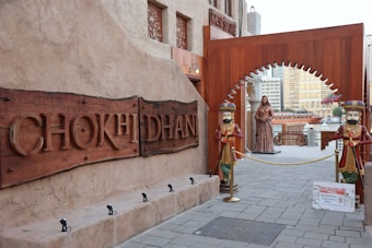 A rustic entrance marked by a large wooden sign displaying the name 'CHOKHI DHANI.' The entrance is flanked by two colorful, traditional statues dressed in ornate outfits. Beyond the archway, there's a statue of a woman and a sign indicating a restricted area for authorized personnel only. The surrounding architecture features earthy tones and intricate designs, typical of traditional Indian styles.