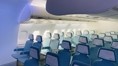 A spacious and well-lit commercial airplane cabin with rows of unoccupied seats. The seats are upholstered in light blue with white headrests, and each has an armrest. Overhead compartments are closed and windows allow natural light to illuminate the interior.