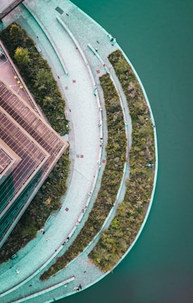 An aerial view of a semicircular, terraced landscape featuring a combination of green spaces and pathways. The terraces are bordered by greenery and surrounded by a body of water, creating a harmonious blend of natural and architectural elements.