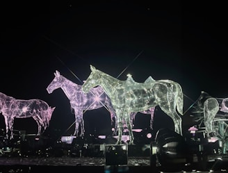 A group of sculptures depicting horses made from a reflective, translucent material that glows in the dark. The sculptures are positioned on a platform with various lights and fixtures around them, creating a futuristic and ethereal atmosphere. The shimmer and sparkle of the material catch the light in complex patterns.