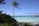 A tranquil tropical beach scene with a tall palm tree leaning slightly toward a turquoise lagoon. Thatched overwater bungalows sit on stilts above the lagoon, with a backdrop of lush greenery and a distant shoreline. The sky is mostly clear, bathed in warm sunlight.