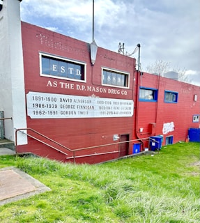 A red brick building features historical signage with the text ESTD. 1889 and the name D.P. Mason Drug Co. Below the windows, a list of names and years is painted onto the wall, providing a timeline of individuals associated with the business. The building is set against a background of grass and a partly cloudy sky. A metal railing leads up the grassy slope towards the building.