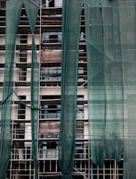 A building under construction is covered with green mesh fabric, with scaffolding visible behind it. The fabric appears to be draped unevenly, allowing glimpses of windows and the building's unfinished surfaces.