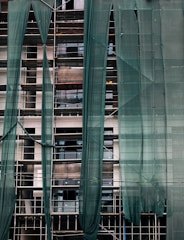 A building under construction is covered with green mesh fabric, with scaffolding visible behind it. The fabric appears to be draped unevenly, allowing glimpses of windows and the building's unfinished surfaces.