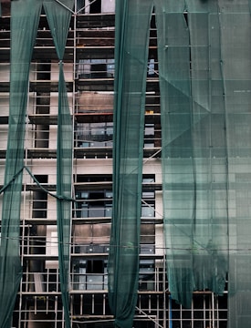 A building under construction is covered with green mesh fabric, with scaffolding visible behind it. The fabric appears to be draped unevenly, allowing glimpses of windows and the building's unfinished surfaces.