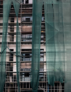 A building under construction is covered with green mesh fabric, with scaffolding visible behind it. The fabric appears to be draped unevenly, allowing glimpses of windows and the building's unfinished surfaces.