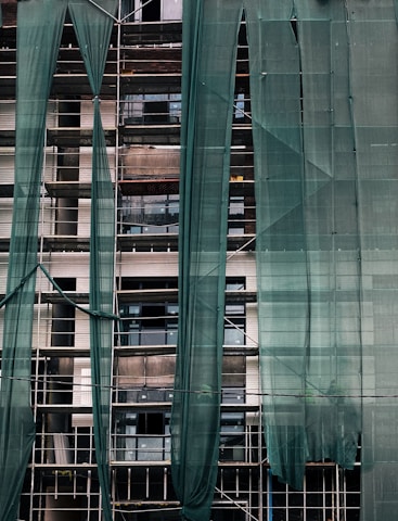 A building under construction is covered with green mesh fabric, with scaffolding visible behind it. The fabric appears to be draped unevenly, allowing glimpses of windows and the building's unfinished surfaces.
