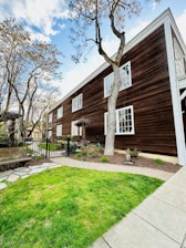 A wooden, two-story building with white-framed windows and a brown exterior is situated amidst a garden area. The foreground features a neatly trimmed green lawn and a stone path leading towards the house. Leafless trees with intricate branches partially obstruct the view of the building. A black metal fence encloses part of the garden, near a wooden bench placed along a paved area.