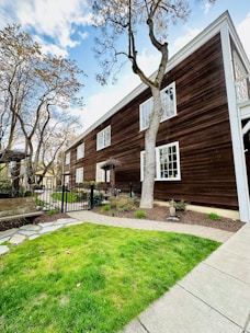 A wooden, two-story building with white-framed windows and a brown exterior is situated amidst a garden area. The foreground features a neatly trimmed green lawn and a stone path leading towards the house. Leafless trees with intricate branches partially obstruct the view of the building. A black metal fence encloses part of the garden, near a wooden bench placed along a paved area.
