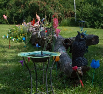 A garden scene with a decorative bird bath in the foreground, surrounded by colorful artificial flowers and other garden decorations. There is a large log positioned horizontally in the center, with various garden ornaments placed on and around it, including a black owl statue and several hanging lanterns. The surrounding grass is green and well-maintained, and there are various plants and flowers in bloom.