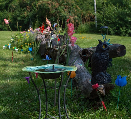 A garden scene with a decorative bird bath in the foreground, surrounded by colorful artificial flowers and other garden decorations. There is a large log positioned horizontally in the center, with various garden ornaments placed on and around it, including a black owl statue and several hanging lanterns. The surrounding grass is green and well-maintained, and there are various plants and flowers in bloom.