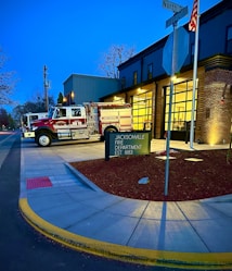 A fire station with a fire truck parked outside on the driveway. The building has brick and metal siding with a visible entrance and large glass garage doors. A street intersection is seen with a sign reading 'Jacksonville Fire Department EST 1883' on a well-maintained sidewalk. An American flag is displayed on a flagpole near the building.