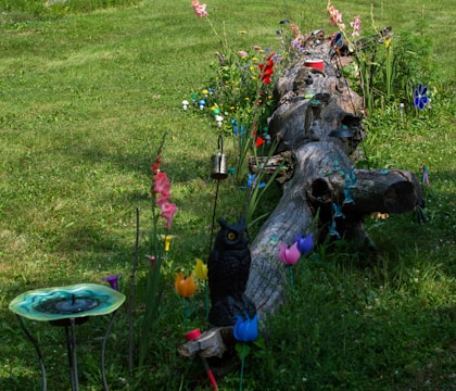A garden scene featuring a large, weathered log surrounded by vibrant flowers and decorations. Various colorful flowers and metal ornamental pieces adorn the area, including an artificial owl figure and a decorative birdbath. The grass is lush and green, providing a natural backdrop for the array of hues and textures.