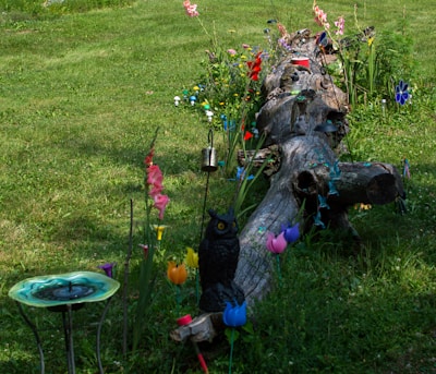 A garden scene featuring a large, weathered log surrounded by vibrant flowers and decorations. Various colorful flowers and metal ornamental pieces adorn the area, including an artificial owl figure and a decorative birdbath. The grass is lush and green, providing a natural backdrop for the array of hues and textures.