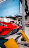 Red sports car on a car lift inside a modern automotive workshop or showroom. The car's sleek design and aerodynamic features are prominently displayed. The environment is clean and well-lit with large windows and multiple other vehicles in the background. Yellow safety barriers are positioned in front of the car.