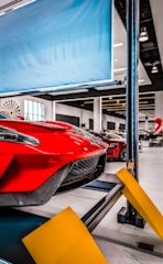 Red sports car on a car lift inside a modern automotive workshop or showroom. The car's sleek design and aerodynamic features are prominently displayed. The environment is clean and well-lit with large windows and multiple other vehicles in the background. Yellow safety barriers are positioned in front of the car.