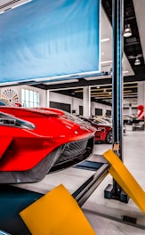 Red sports car on a car lift inside a modern automotive workshop or showroom. The car's sleek design and aerodynamic features are prominently displayed. The environment is clean and well-lit with large windows and multiple other vehicles in the background. Yellow safety barriers are positioned in front of the car.