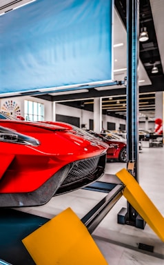 Red sports car on a car lift inside a modern automotive workshop or showroom. The car's sleek design and aerodynamic features are prominently displayed. The environment is clean and well-lit with large windows and multiple other vehicles in the background. Yellow safety barriers are positioned in front of the car.