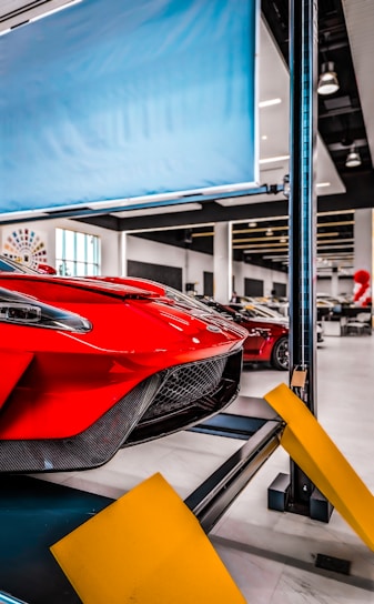 Red sports car on a car lift inside a modern automotive workshop or showroom. The car's sleek design and aerodynamic features are prominently displayed. The environment is clean and well-lit with large windows and multiple other vehicles in the background. Yellow safety barriers are positioned in front of the car.