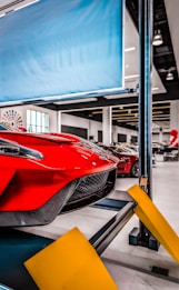 Red sports car on a car lift inside a modern automotive workshop or showroom. The car's sleek design and aerodynamic features are prominently displayed. The environment is clean and well-lit with large windows and multiple other vehicles in the background. Yellow safety barriers are positioned in front of the car.