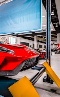 Red sports car on a car lift inside a modern automotive workshop or showroom. The car's sleek design and aerodynamic features are prominently displayed. The environment is clean and well-lit with large windows and multiple other vehicles in the background. Yellow safety barriers are positioned in front of the car.
