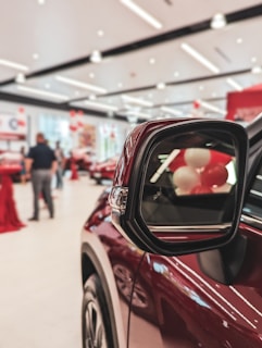 A close-up view of a maroon-colored car's side mirror in focus. The background shows a blurred interior of a showroom with people, red and white balloons, and other cars.