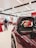 A close-up view of a maroon-colored car's side mirror in focus. The background shows a blurred interior of a showroom with people, red and white balloons, and other cars.