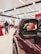 A close-up view of a maroon-colored car's side mirror in focus. The background shows a blurred interior of a showroom with people, red and white balloons, and other cars.