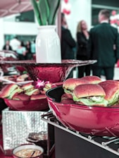 A table is set with red bowls containing sliders or small sandwiches. In the background, a group of people in formal attire appear to be engaged in conversation, with a vase and decorative elements enhancing the scene.