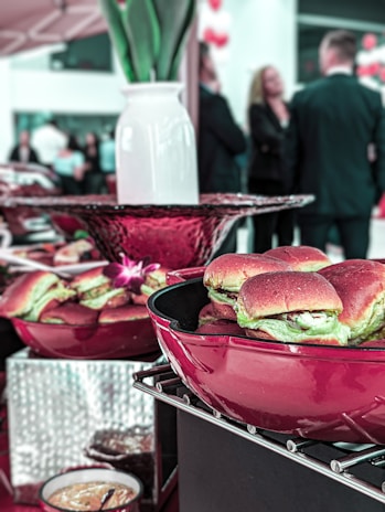 A table is set with red bowls containing sliders or small sandwiches. In the background, a group of people in formal attire appear to be engaged in conversation, with a vase and decorative elements enhancing the scene.