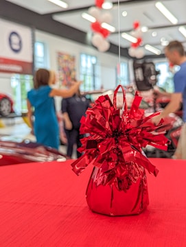 In the foreground, a shiny red foil decoration with ribbons is placed on a red table. The background shows a blurred indoor setting with people engaged in conversation, decorative balloons, and what appears to be cars on display.
