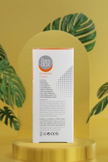 A box of Paradox sunscreen cream is prominently displayed. The packaging is white with orange and gray accents, featuring text detailing its features such as oil-free, anti-aging, and high protection. The box is placed against a soft golden background with stylized green leaves, adding a natural and elegant touch.