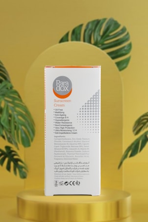 A box of Paradox sunscreen cream is prominently displayed. The packaging is white with orange and gray accents, featuring text detailing its features such as oil-free, anti-aging, and high protection. The box is placed against a soft golden background with stylized green leaves, adding a natural and elegant touch.