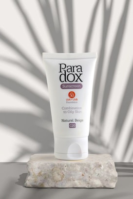 A tube of Paradox sunscreen stands upright on a textured stone surface. Its label indicates SPF 50 protection against UVA/UVB rays, suitable for combination to oily skin, with a natural beige tone denoted as C20. Shadows of plants or leaves are artfully cast on the background, adding a natural element to the composition.
