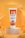 A tube of Dermalift Sunlift tinted sunscreen cream with SPF 50 is displayed. The tube is placed on a marble-like pedestal and has a vibrant orange and white design. The background features tropical palm leaves and an orange gradient, creating a warm, summer ambiance.