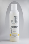 A bottle of Delano brand shampoo, labeled for colored and damaged hair with Argan oil. The design includes a botanical illustration and the text 'Sulfate Free'. The bottle is primarily white with gray and yellow accents.