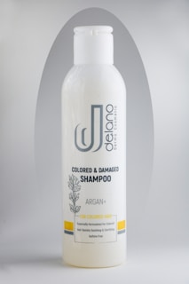 A bottle of Delano brand shampoo, labeled for colored and damaged hair with Argan oil. The design includes a botanical illustration and the text 'Sulfate Free'. The bottle is primarily white with gray and yellow accents.