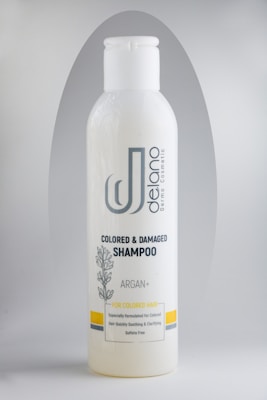A bottle of Delano brand shampoo, labeled for colored and damaged hair with Argan oil. The design includes a botanical illustration and the text 'Sulfate Free'. The bottle is primarily white with gray and yellow accents.