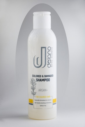 A bottle of Delano brand shampoo, labeled for colored and damaged hair with Argan oil. The design includes a botanical illustration and the text 'Sulfate Free'. The bottle is primarily white with gray and yellow accents.